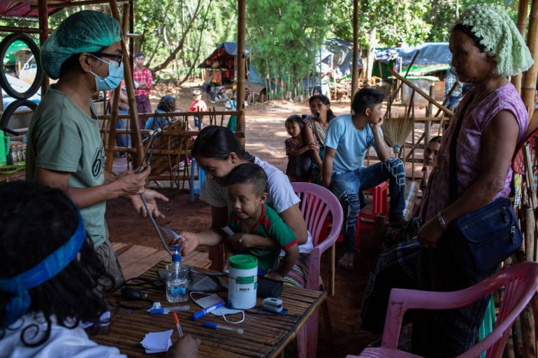 Displaced people seek medical treatment from a volunteer team. A woman is seated at a pink plastic chair at a table in a bamboo building. A medic is speaking to another woman. They are wearing a hairnet and face mask.