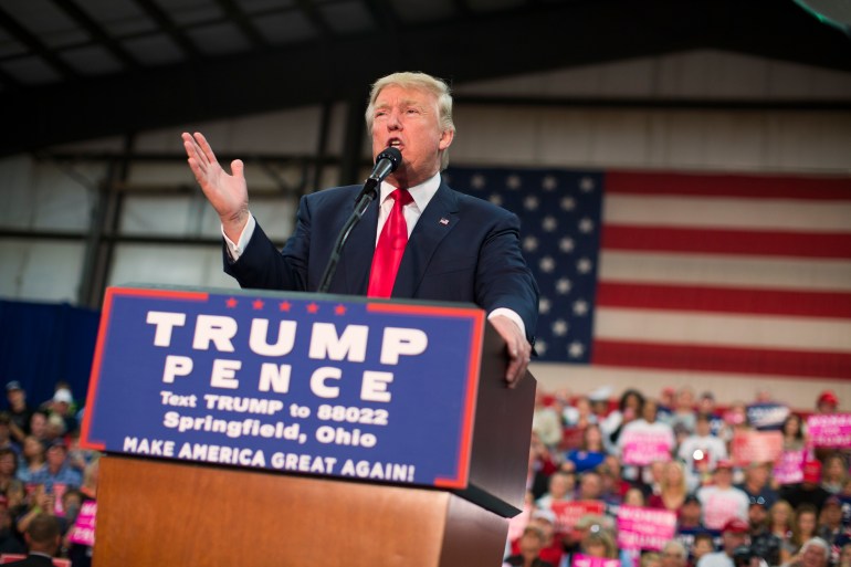 Donald Trump stands at a podium in his blue suit and red tie, lifting one hand in gesture. Behind him is a massive US flag, and the podium is decorated with the text: "Trump-Pence. Text Trump to 8802. Springfield, Ohio. Make America Great Again."