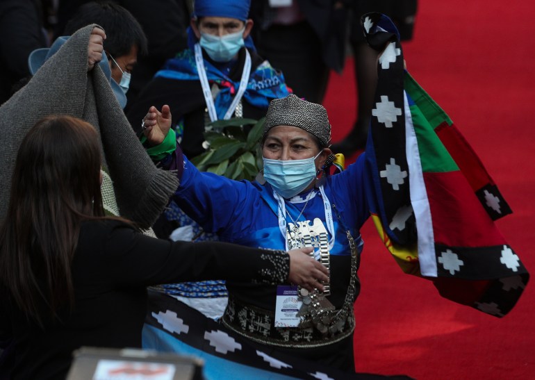 A Mapuche representative raises her arms as she celebrates her election to the constitutional council in 2022. A red carpet is visible behind her, and in one hand she carries a flag.