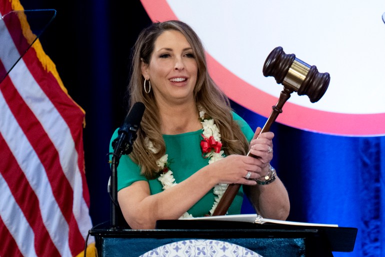 Ronna McDaniel, wearing a green shirt and a garland of flowers around her neck and carrying an oversized gavel, stands at a podium in California.