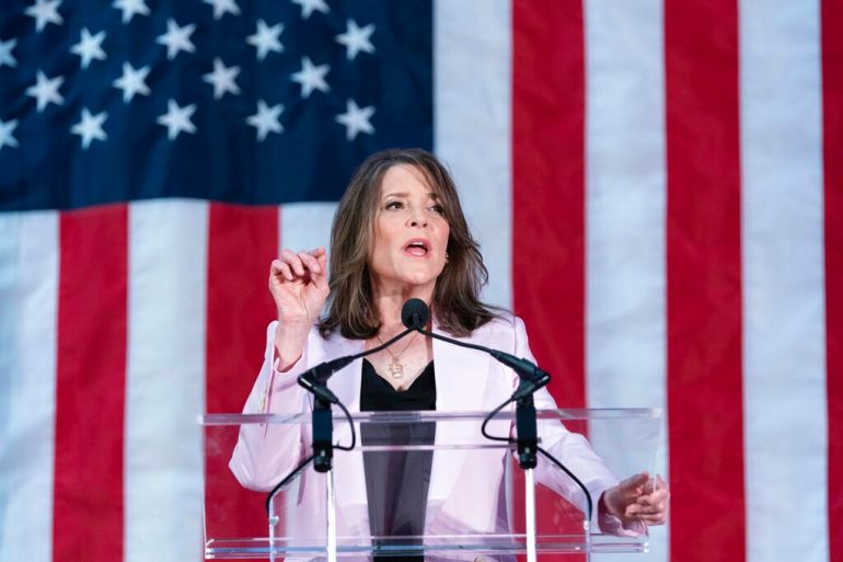 Marianne Williamson speaking on a podium in front of a large US flag