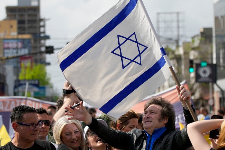 Javier Milei, ao ar livre no meio de uma multidão, segura uma bandeira israelense.