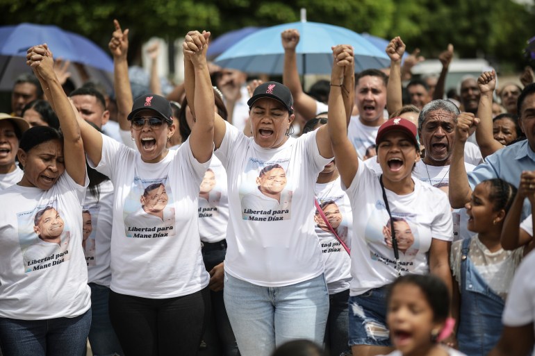 Frauen mit Ballkappen und T-Shirts fassen sich an den Händen und heben ihre Arme in die Luft, während sie protestieren, um auf die Freilassung von Luis Diaz‘ Vater zu drängen.  Auf jedem ihrer T-Shirts ist das Gesicht des Vaters aufgedruckt.