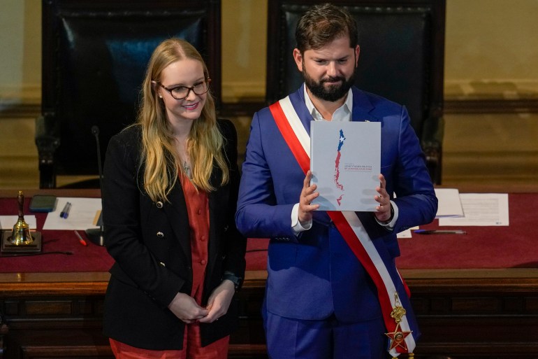 Gabriel Boric, wearing a blue suit and red-white-and-blue sash, holds up a copy of the draft constitution next to Beatriz Hevia, the president of the Constitutional Council.