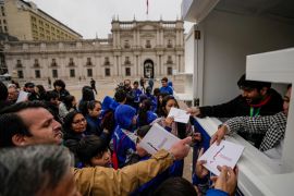 A crowd of people gather in front of the window of a booth in Santiago, where people hand out copies of the latest constitutional draft. The presidential palace is visible in the background.