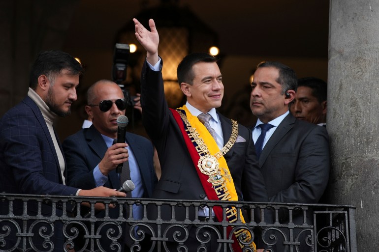 Daniel Noboa stands on a balcony with officials in dark suits, wearing a sash in the colors of Ecuador's flag and waving to the crowds below.