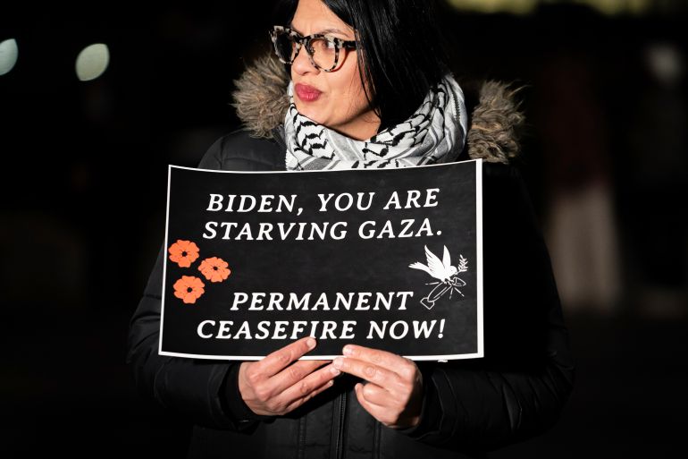 Congresswoman Rashida Tlaib stands against a night sky, holding up a sign that reads, "Biden you are starving Gaza. Permanent ceasefire now."