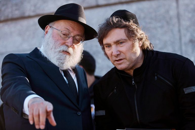 Javier Milei, wearing a black yarmulke, speaks to a rabbi who points to something in front of them.