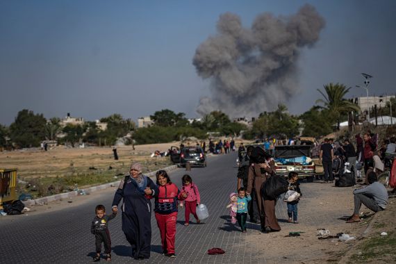 a family carrying their belongings walk in the foreground as a a black plume of smoke can be seen behind them
