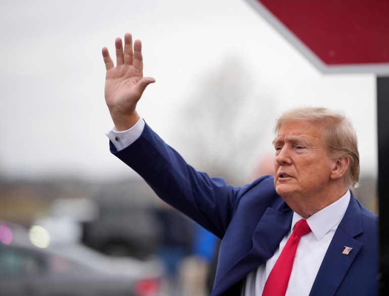 Donald Trump, dressed in a blue suit and red tie, lifts his arm in the air to wave against a grey sky.
