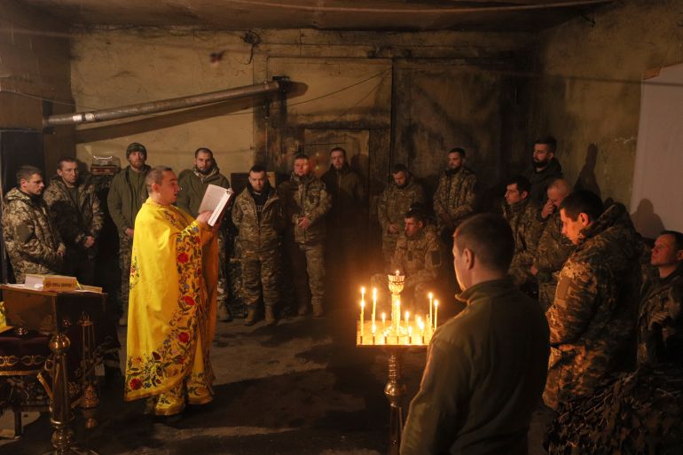 Soldados ucranianos ouvindo um capelão recitar uma oração.  Eles estão na escuridão, exceto pelas velas
