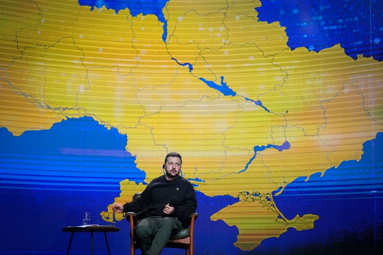 Volodymyr Zelenskyy sitting on stage during his annual press conference. A map of Ukraine is in blue and yellow behind him.