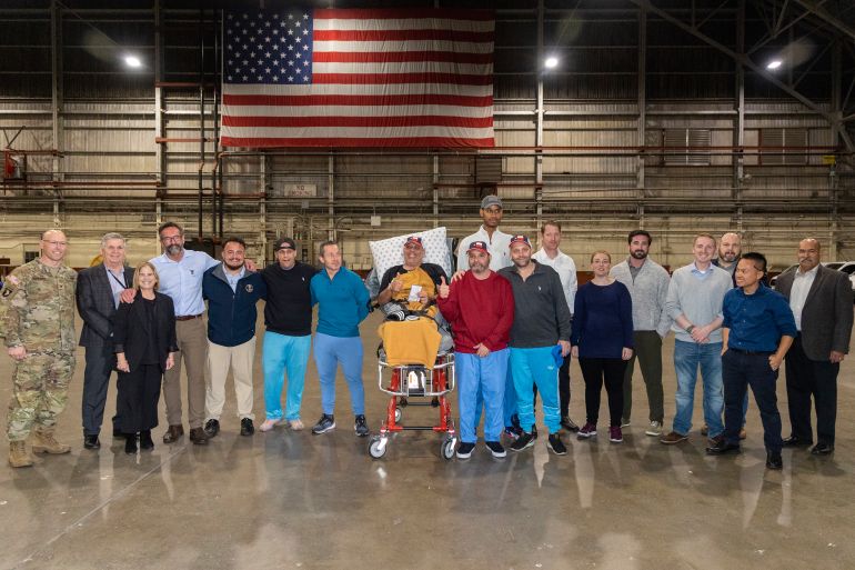 A group photo of the freed Americans in an aircraft hangar at the base in San Antonio. There's a US flag behind them. Government officials are also in the photo