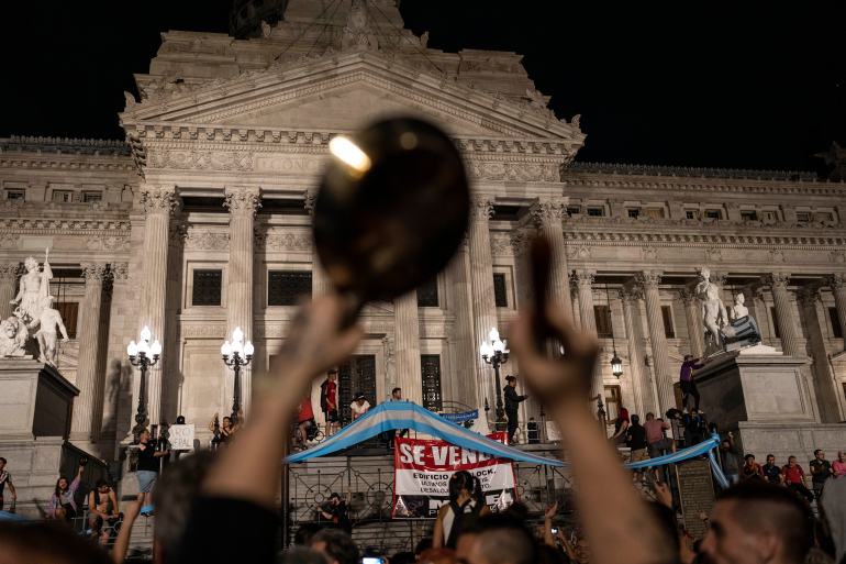 A protester lifts their arms high into the air to bang pots and pans in front of Argentina's stately National Congress, a neoclassical building with columns. Around the protester, others demonstrate as well, with banners and Argentine flags.