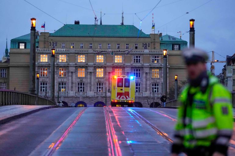 An ambulance with its lights flashing heading towards the Charles University. A police officer is in front. The building is behind.