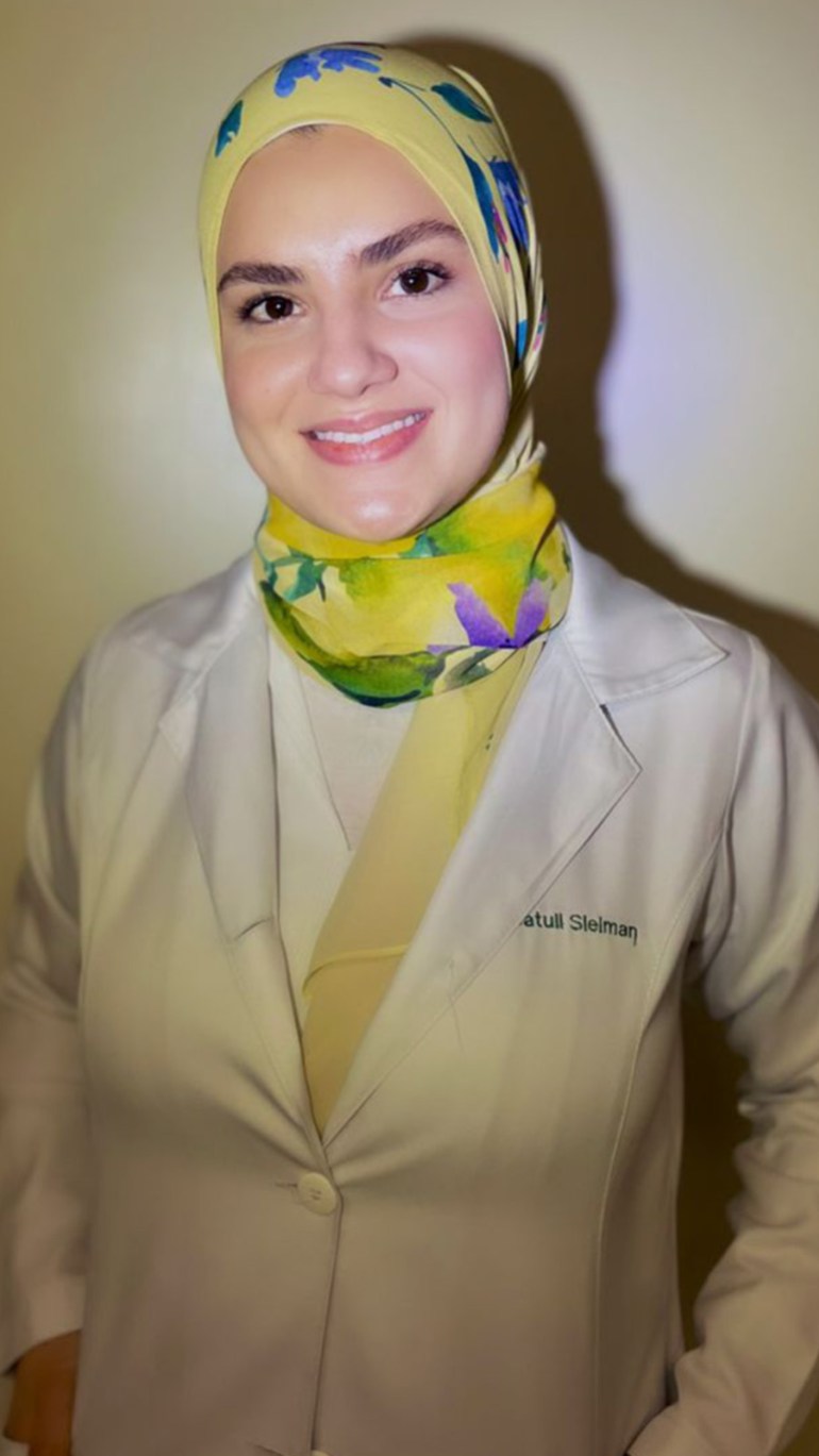 A woman smiles in a portrait photo. She wears a white doctor's coat and a yellow patterned hijab.