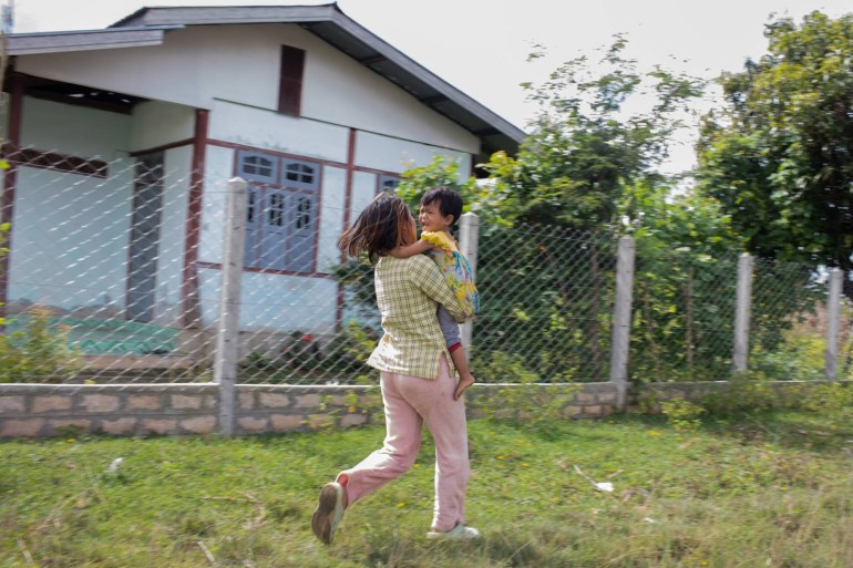 A woman running with a young child in her arms. There is a house behind them. They look scared.