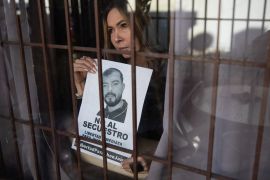 A woman holds up a missing person's poster with her brother's face on it, as she stares through a window protected with metal bars.
