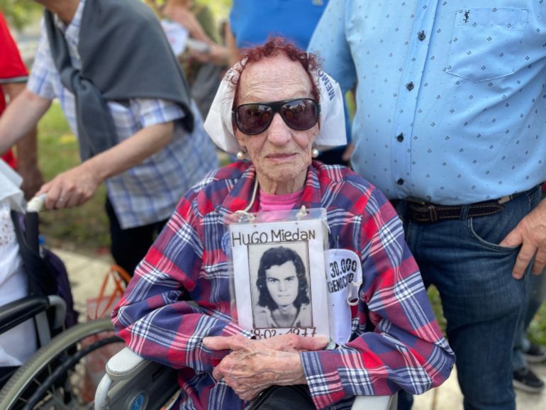 Elia Espin, a member of the Grandmothers of the Plaza de Mayo — a group of women who demonstrate for the return of people disappeared during the dictatorship — holds up an old black-and-white photo of her young son Hugo Miedan, who was among those abducted. She sits in a wheelchair, wearing a plaid shirt, sunglasses and a white scarf in her hair.