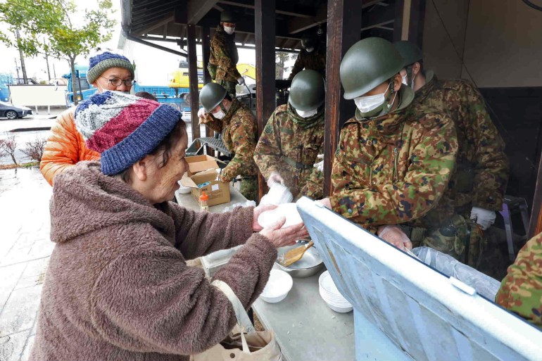 Japanese soldiers preparing hot meals for quake survivors at an outdoor kitchen. An elderly woman in a coat nd woolly hat is taking some of the food.