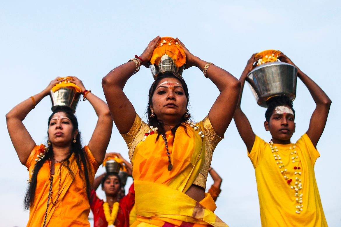 Two women and a young man carrying pots of milk on their heads. They are dressed in orangey/yellow clothing.