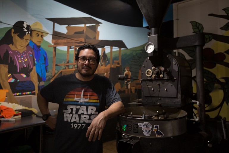 Um homem com uma camiseta de Star Wars dos anos 1970 está em uma sala com uma máquina de metal escuro – provavelmente para torrar café – e um mural atrás dele.