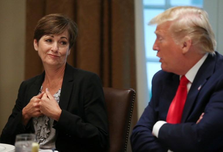 Kim Reynolds, gesturing with both hands on her chest, sits next to a cross-armed Donald Trump at a table at the White House.