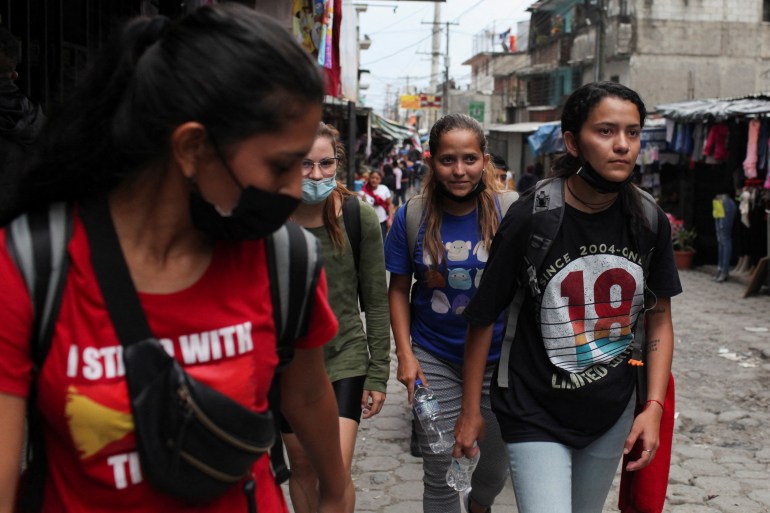 Um grupo de jovens caminha por uma movimentada rua da Guatemala.  Todos usam mochilas e camisetas, com os cabelos puxados para trás e máscaras faciais cobrindo o nariz ou puxadas para baixo do queixo.