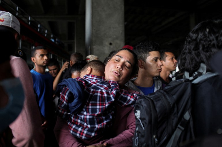 A woman carries her son at night in a crowded station in Guatemala City, a concrete pillar visible behind them.
