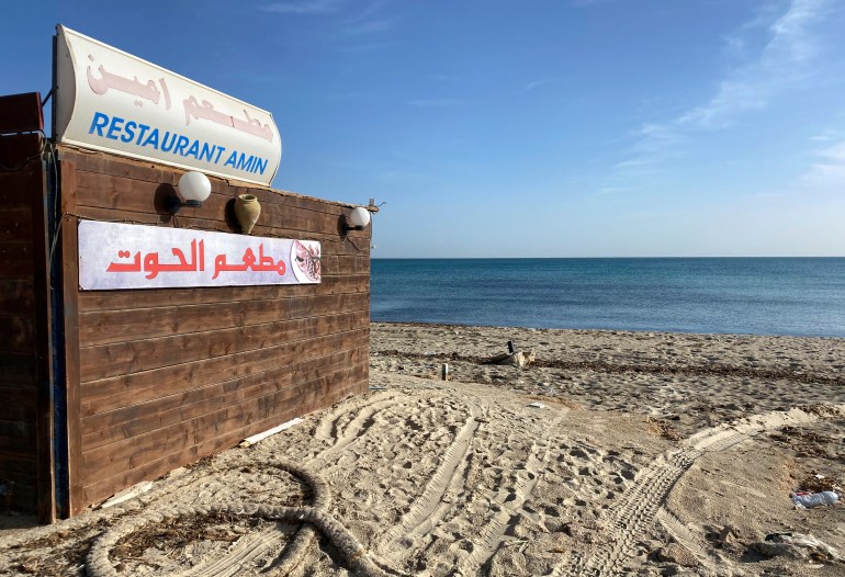 A small wooden restaurant sits on a sandy beach in Zarzis, Tunis, below a clear blue sky.