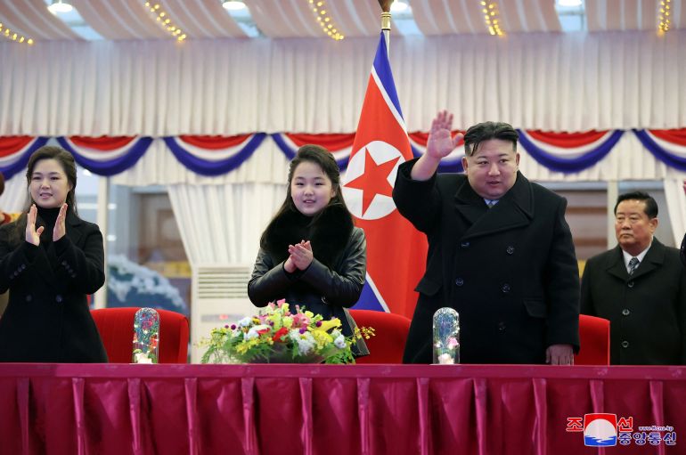 North Korea's Kim Jong Un acknowledging the crowd at a new year's event. He's standing alongside his daughter and wife who are clapping. There is a North Korean flag behind them. They are at a table draped in red cloth with a flower arrangement in the middie