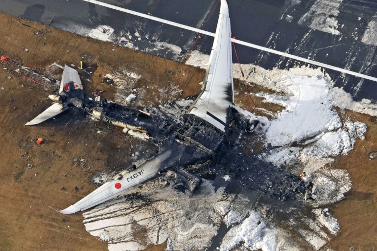 The burnt out wreckage of the JAL plane on the runway. The cabin in the middle is black and destroyed. The wings are still visible and white.