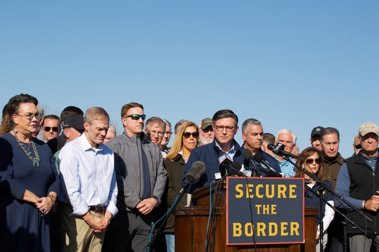Congressional representatives stand behind House Speaker Mike Johnson as he speaks at a podium decorated with a sign that reads, "Secure the border."