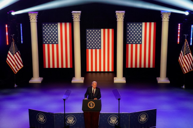 Joe Biden stands at a podium with three US flags hanging between pillars behind him.