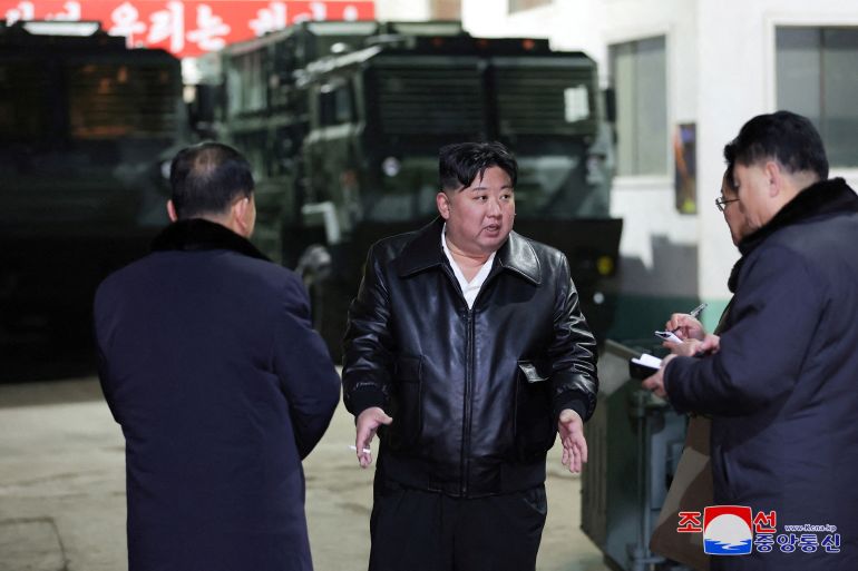 Kim Jong Un em uma fábrica de equipamentos militares.  Ele está vestindo um casaco de couro preto e conversando com autoridades.  Um veículo militar está ao fundo
