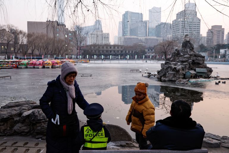 Older people with children at a park in Beijing. Parts of the lake are frozen. Everyone's wearing thick coats. One of the children is wearing a high-vis jacket reading POLICE