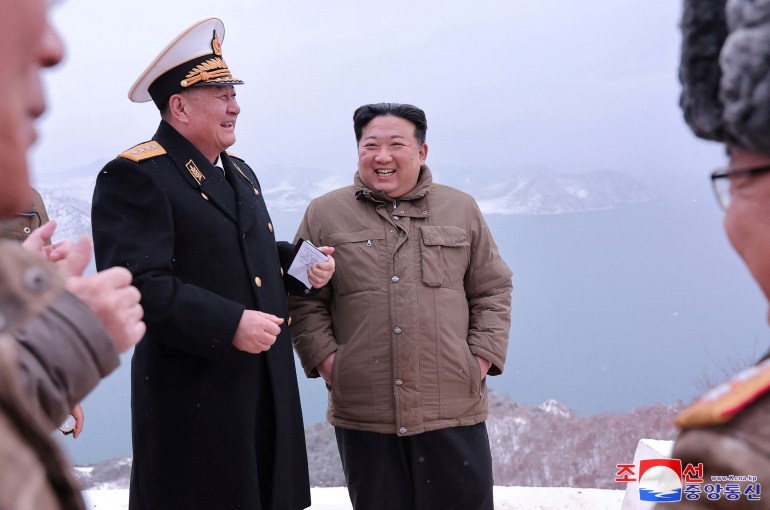 Kim Jong Un lächelt, während er mit Militärbeamten spricht.  Es sieht kalt aus.  Er hat seine Hände in den Taschen.