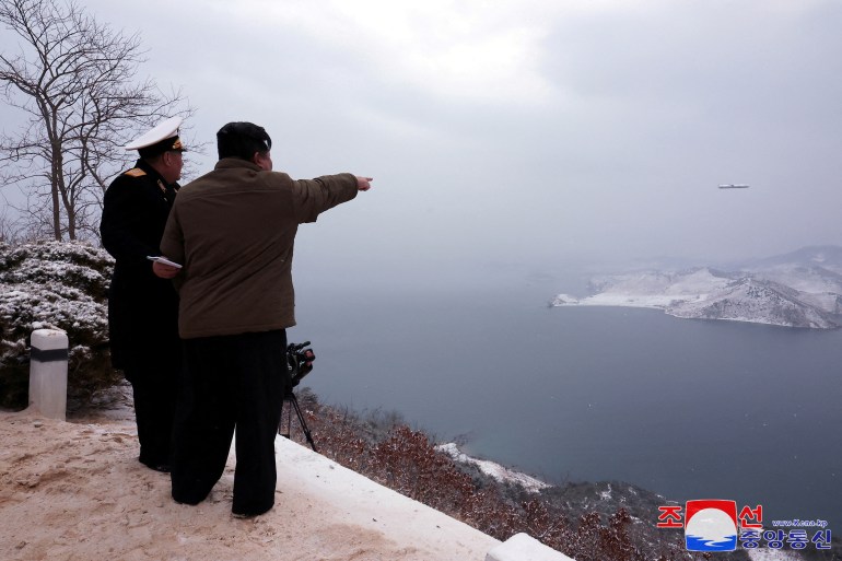 Kim Jong Un pointing at a missile in the sky. He is standing with a man in uniform. There is snow on the ground and they're on a hill looking out to sea