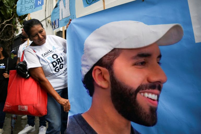 A woman holds up a gigantic poster depicting Nayib Bukele in a white ballcap.