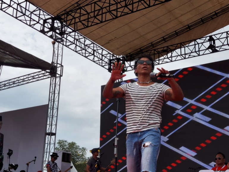 Kaka, the leader of Slank.  He performs on stage.  He wears a black and white striped shirt, jeans and sunglasses.