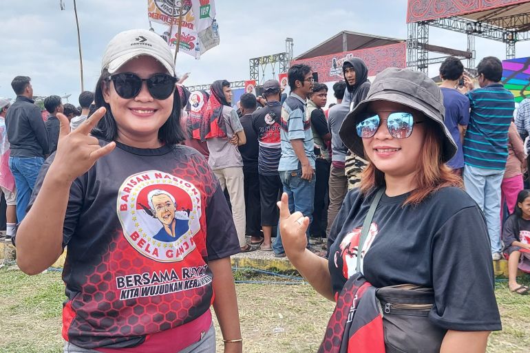 Market sellers Ratna (left) and Lisa (right) are fans of Slank and support Ganjar. Ranta is wearing a T-shirt with Ganjar's face. The stage is behind them. Some people are waving flags.