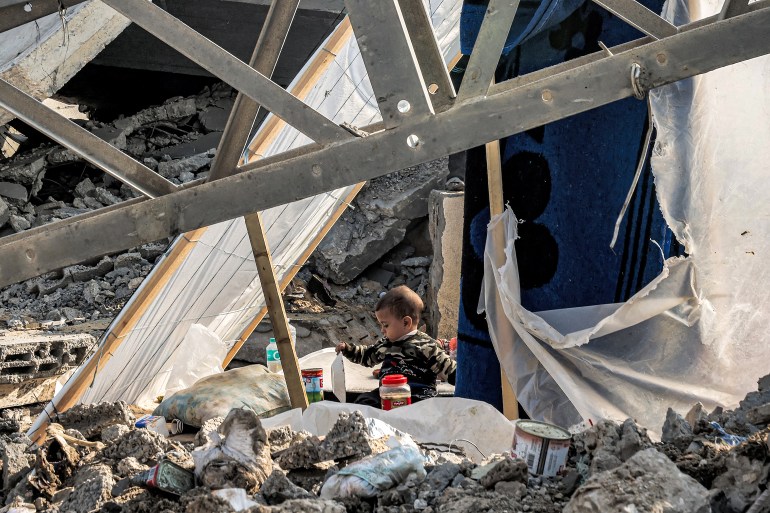 A child holds a plate while sitting between rubble in Rafah