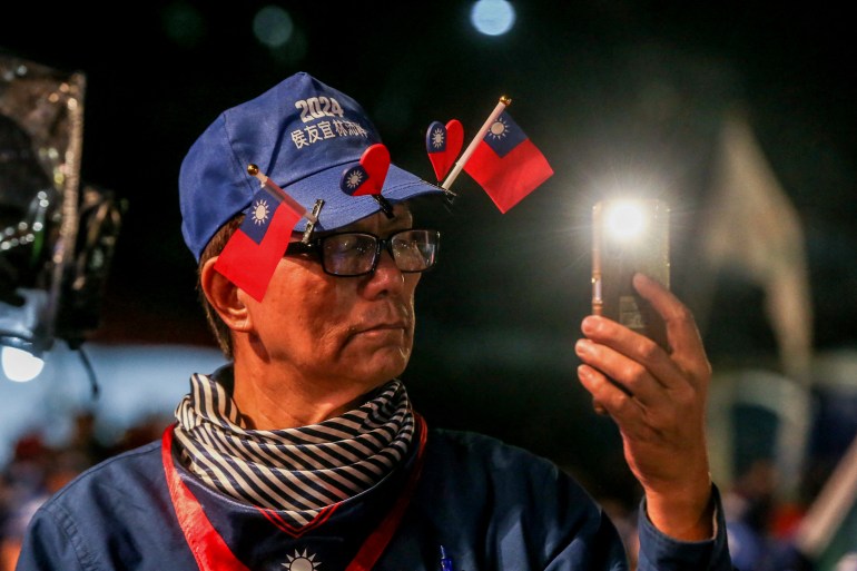 Um apoiador do KMT em um comício.  Ele é um homem mais velho e usa um boné azul decorado com pequenas bandeiras e corações de Taiwan. 