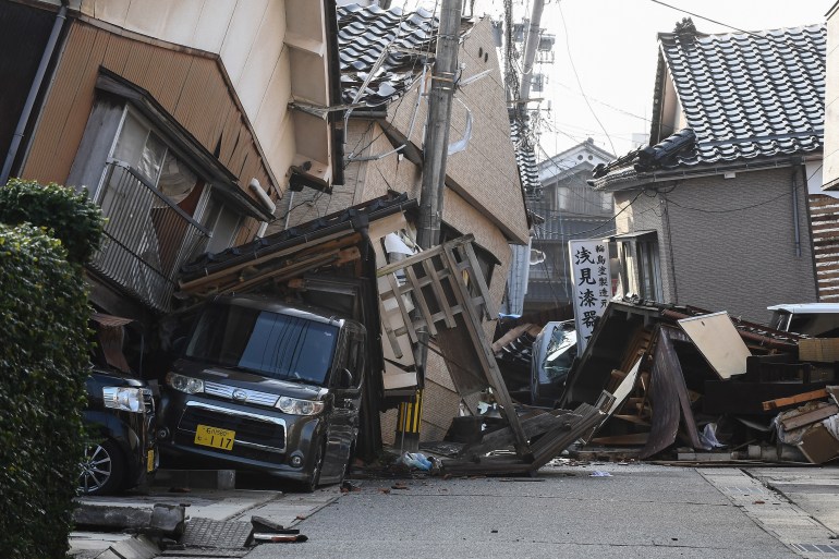 A road blocked by collapsed houses. Cars are also crushed, and pylons are at an angle.