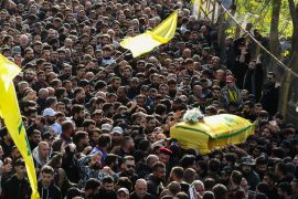 A big crowd with two yellow Hezbollah flags waving and a coffin draped in Hezbollah's yellow flag carried by the mourners in the crowd.