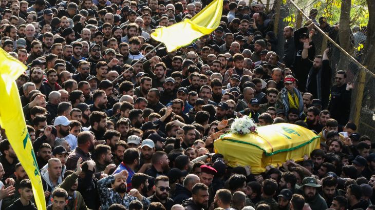 A big crowd with two yellow Hezbollah flags waving and a coffin draped in Hezbollah's yellow flag carried by the mourners in the crowd.