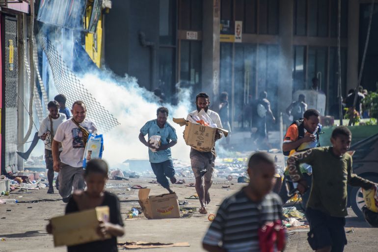 Two men running with looted goods on the streets of Port Moresby. There is smoke behind them and debris on the street.