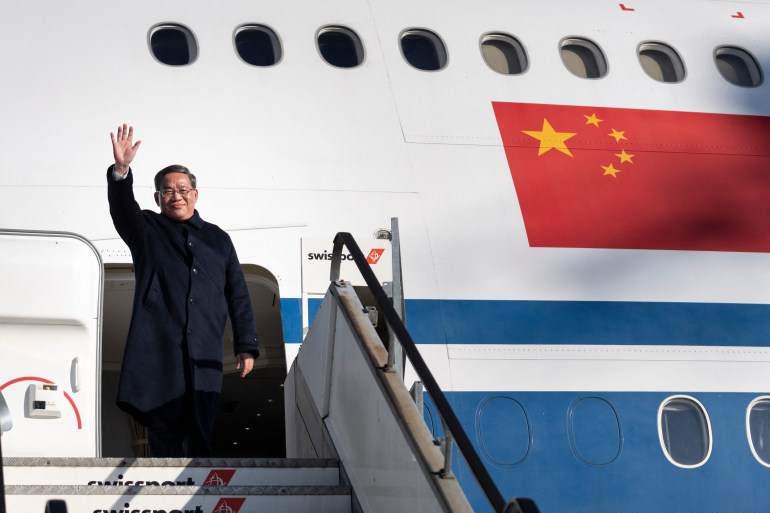 Li Qiang at the top of the steps as he gets off an Air China plane in Zurich. He is wearing a black coat and waving.