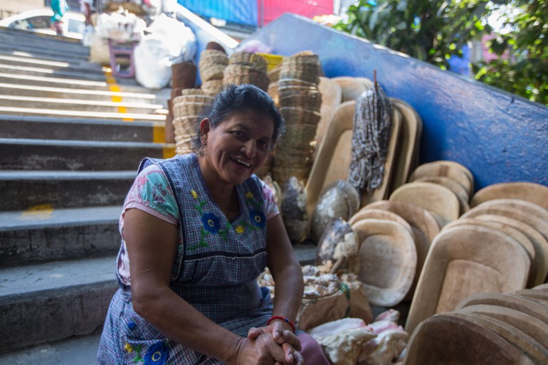 Uma mulher com um vestido estampado está sentada em alguns degraus ao ar livre, ao lado de fileiras de cestos de tecido complexo à venda.