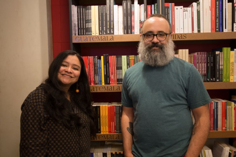 Two people stand side by side in front of a book shelf: a man and a woman, the former wearing a grey T-shirt and the latter wearing black.